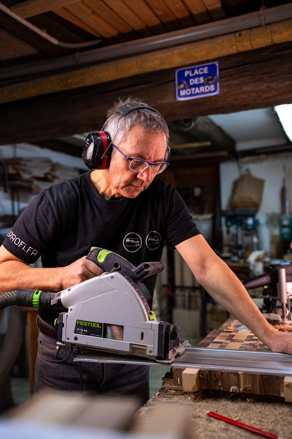 Un homme portant des lunettes et des protections auditives utilise une scie Festool électrique pour couper une planche en bois dans un atelier.